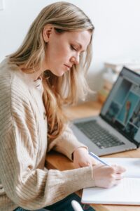 Side view of crop focused female taking notes in planner at table with netbook on blurred background